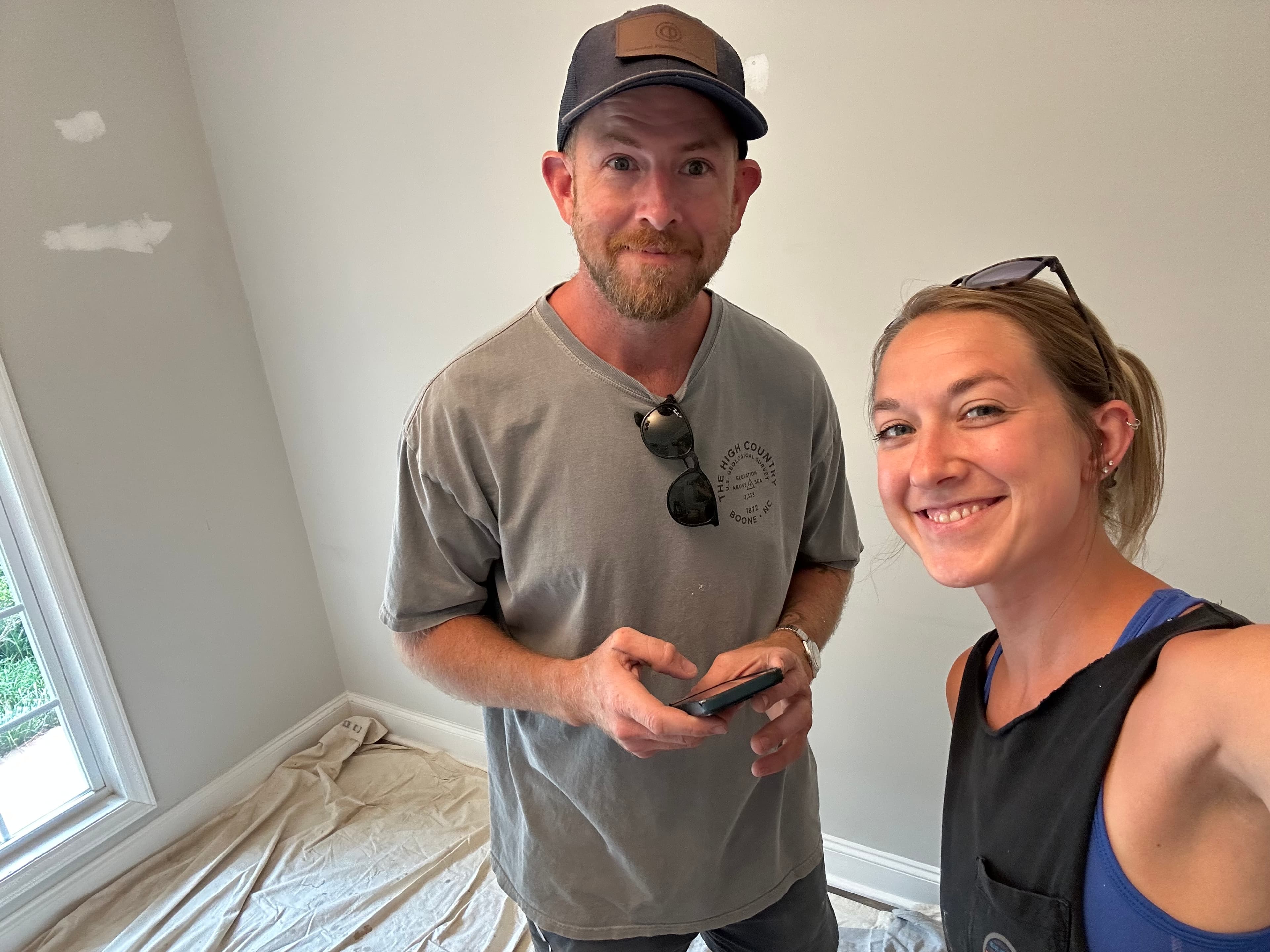 Smiling man and woman stand in a room with drop cloths and patched grey walls.