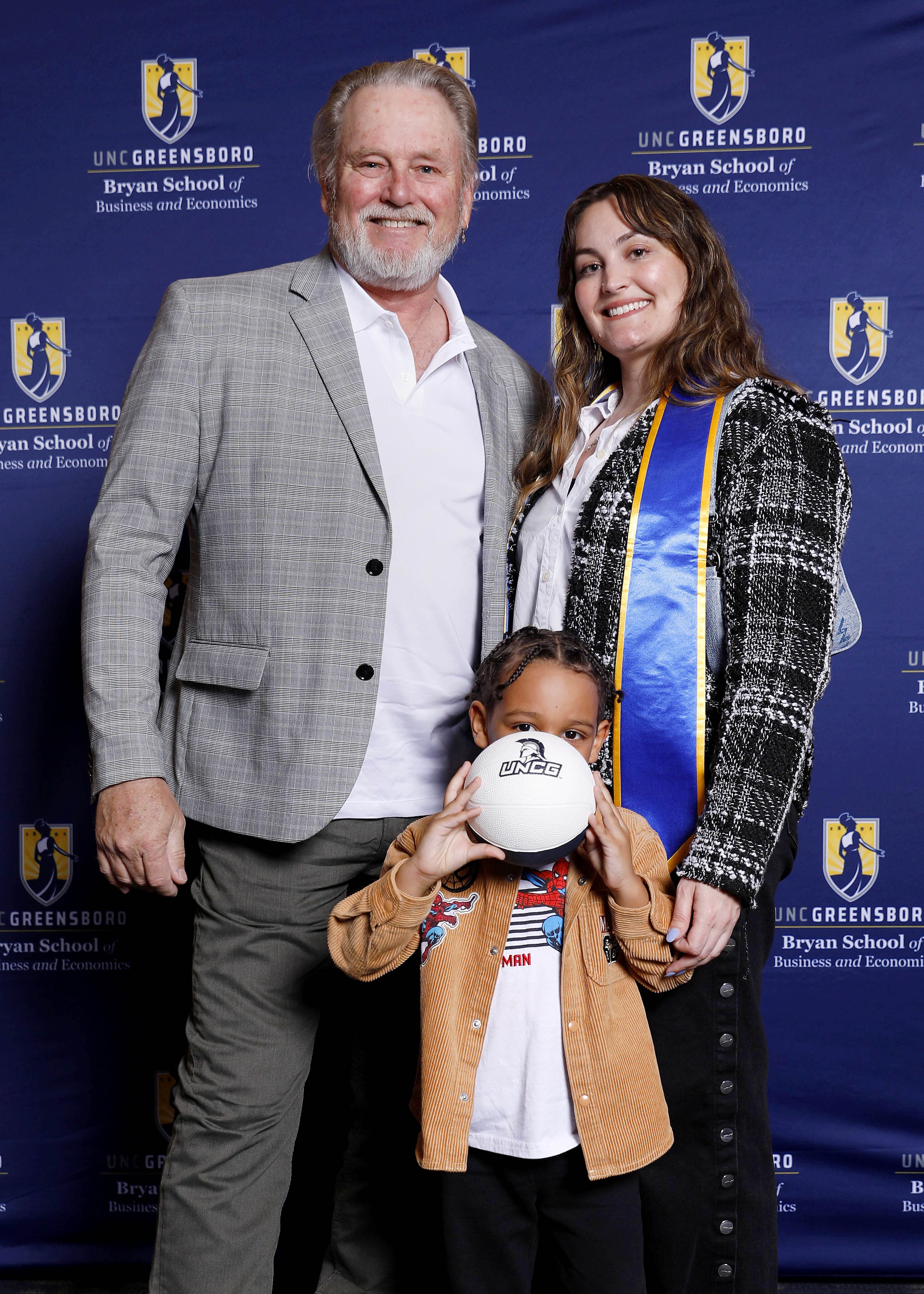 Man, graduate in a sash, and child holding a basketball pose against a UNCG backdrop.
