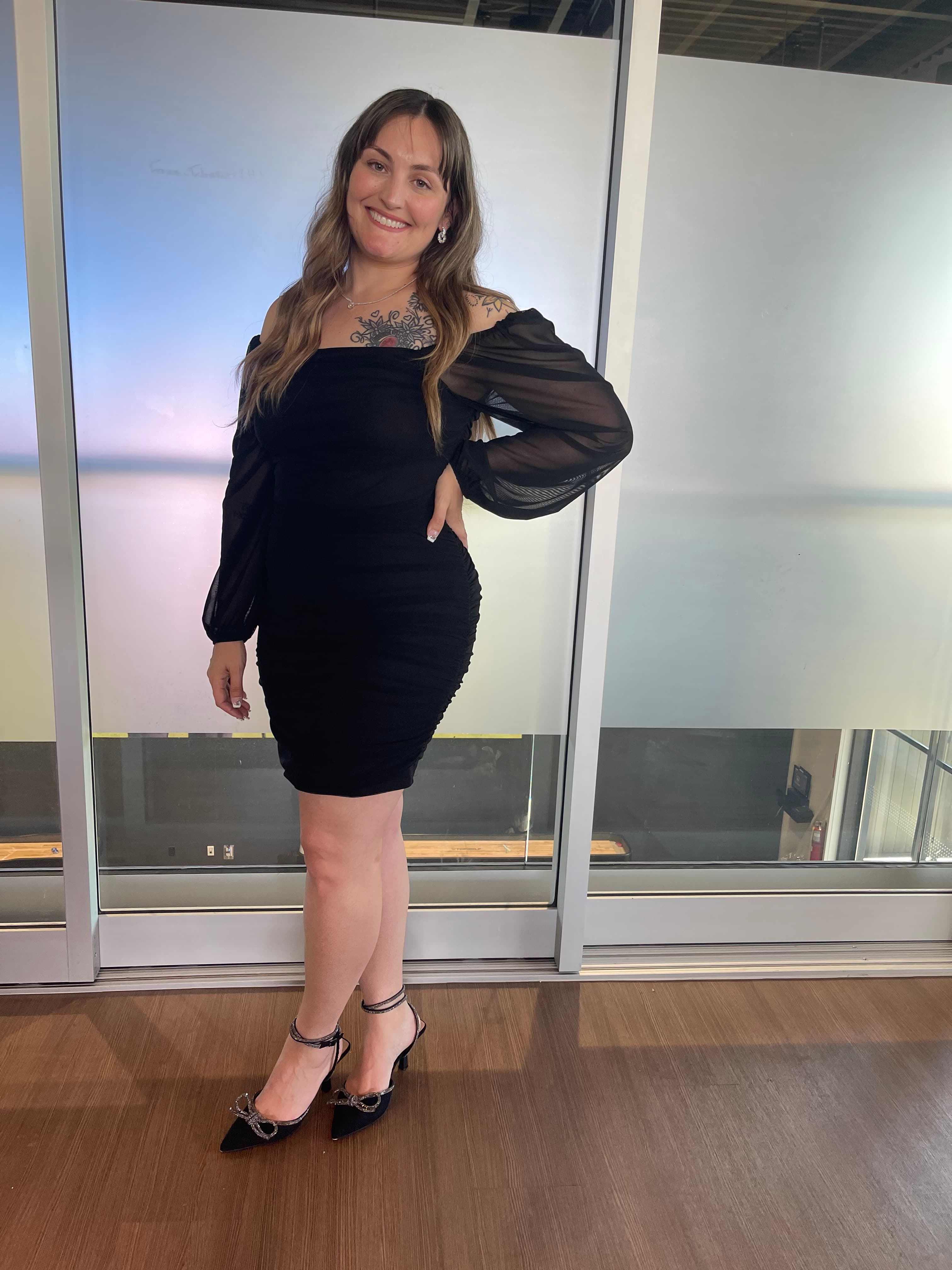 Smiling woman with tattoos wearing a black off-the-shoulder dress and sparkly bow heels.